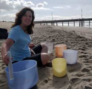 Woman playing singing bowls on sandy beach.